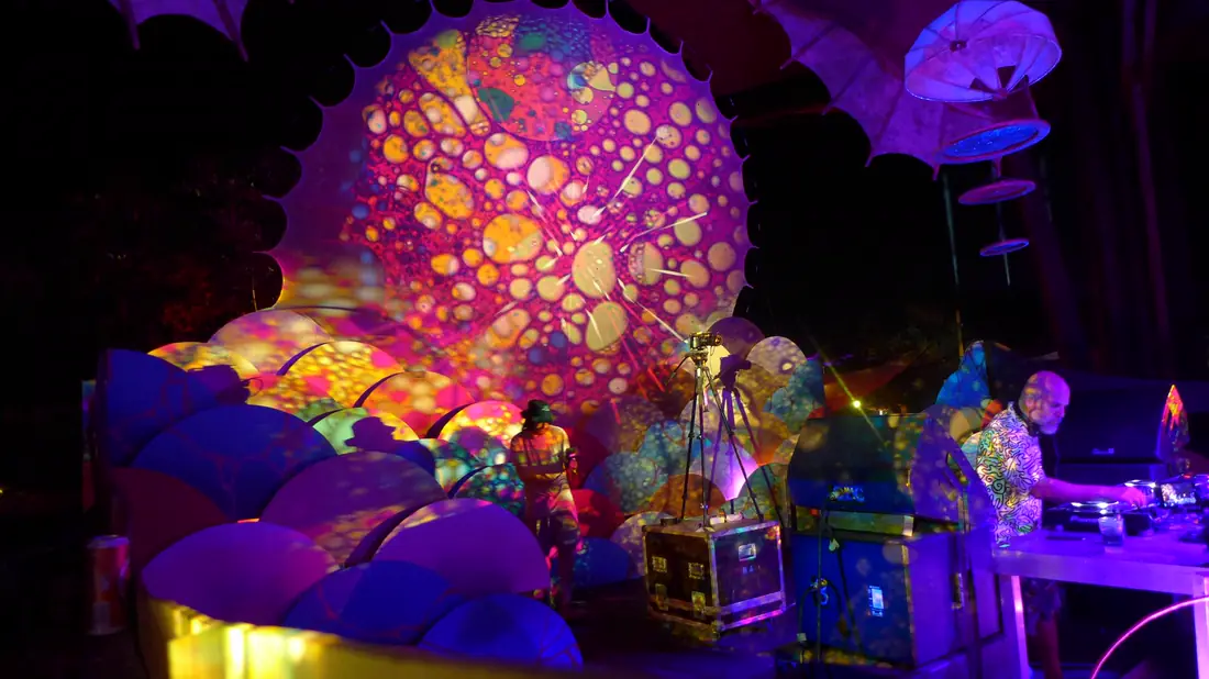 Cúpula de tecido iluminada por padrões de bolhas rosa e amarelas, acima de uma cabine de DJ.
