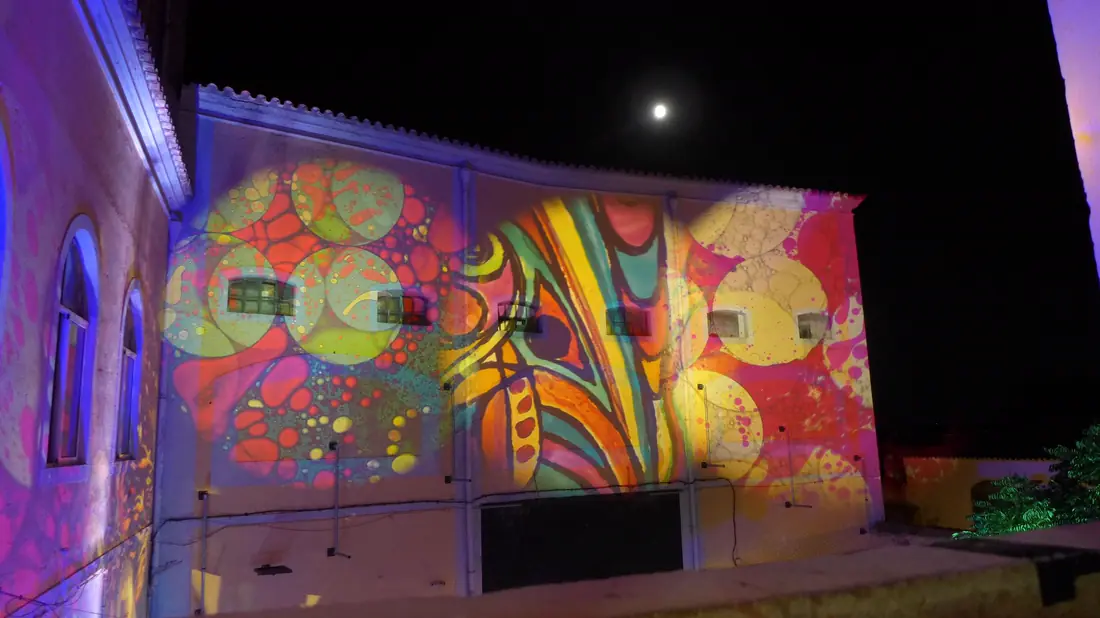 Fachada de um edifício branco inteiramente coberta por projeções coloridas, à noite, com a lua no céu.