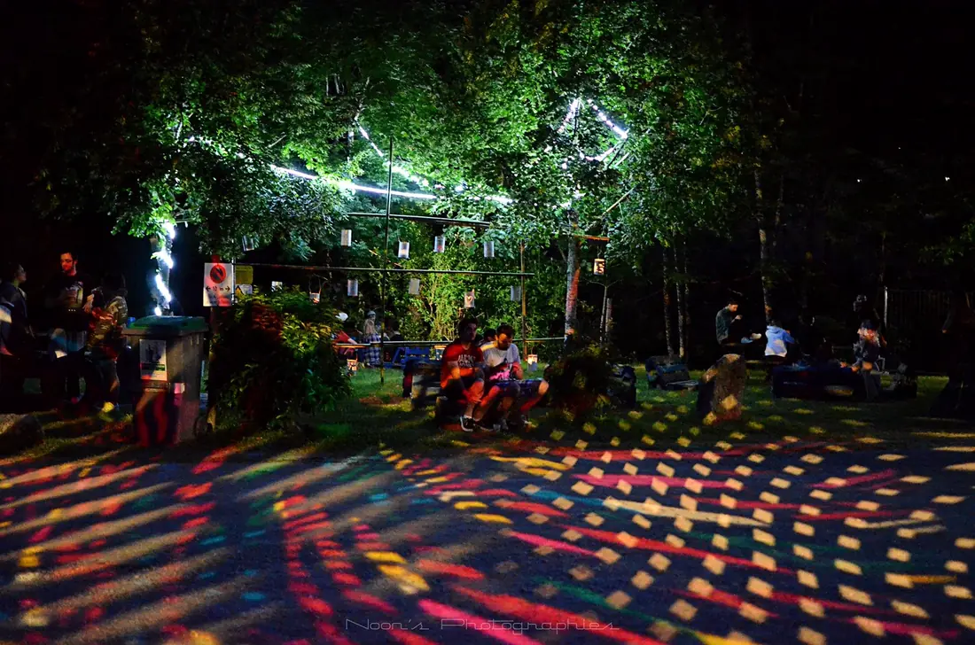 Floresta à noite: o chão de terra está coberto de motivos coloridos projetados, com pessoas sentadas entre as árvores.