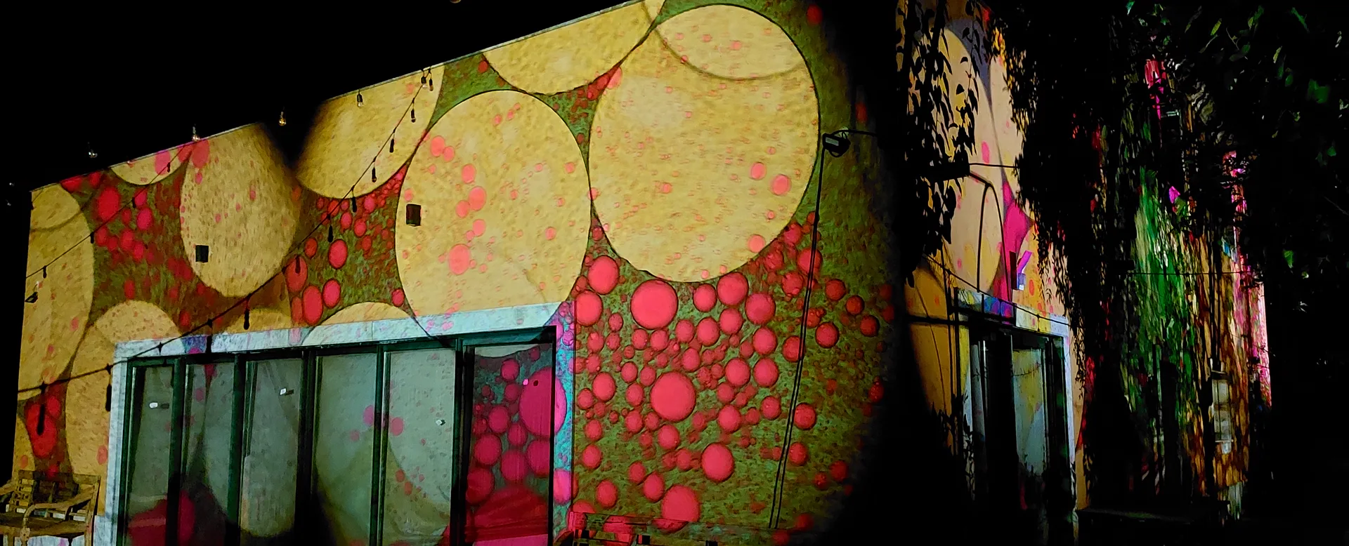 Jardim de um restaurante à noite: ao fundo, a fachada e as portas envidraçadas estão cobertas por projeções coloridas; à frente, mesas e arbustos na penumbra.